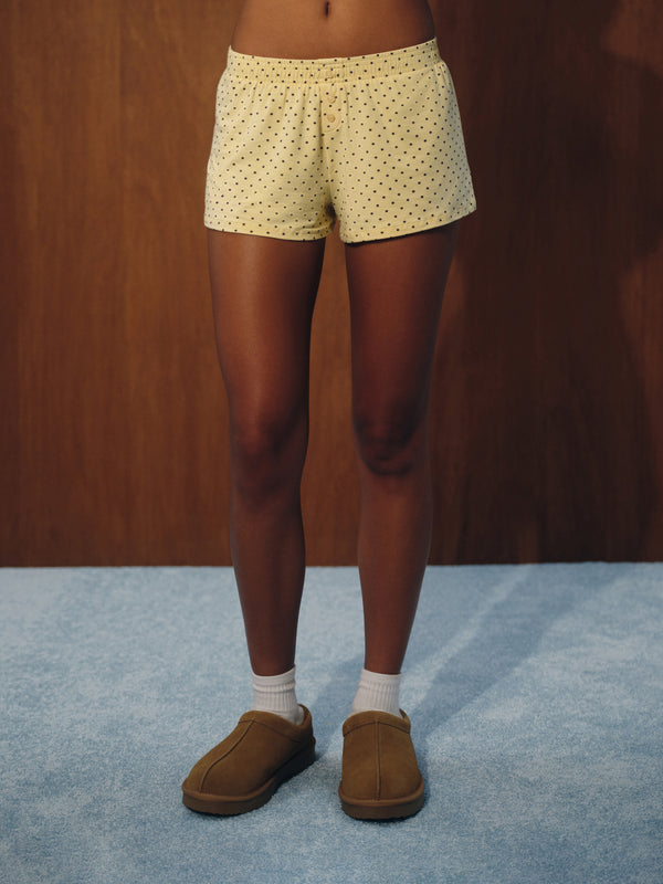 A person from the waist down wearing light yellow shorts with a small black polka dot pattern, white socks, and brown slip-on slippers, standing on a blue carpet against a dark wood panel background.