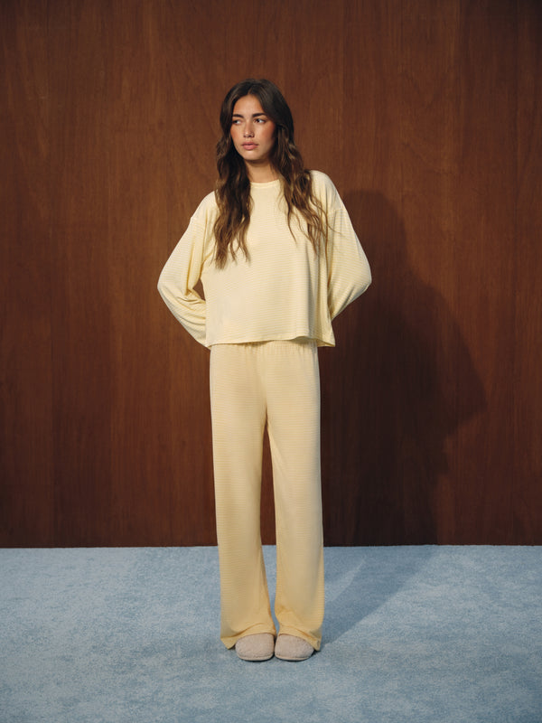A woman stands against a dark wood-paneled wall wearing a matching cream-colored lounge set consisting of a cropped long-sleeve top and wide-leg pants, paired with beige slip-on shoes. 