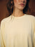 A close-up of a woman wearing a pale yellow long-sleeve top with thin horizontal white stripes against a warm brown background.
