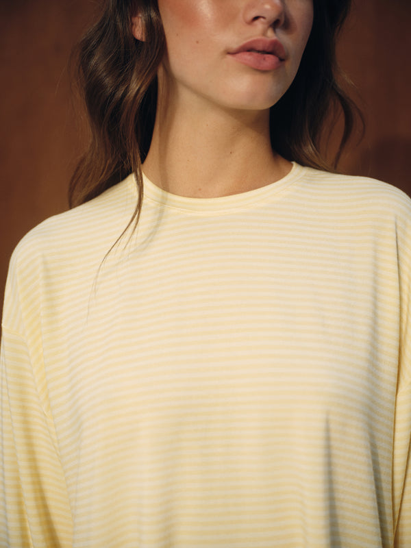 A close-up of a woman wearing a pale yellow long-sleeve top with thin horizontal white stripes against a warm brown background.