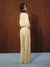A woman stands in profile wearing a pale cream two-piece outfit with a cropped long-sleeve top and wide-leg pants, posed against a dark wood-paneled wall on a light blue floor.