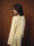 A woman with dark shoulder-length hair is shown from the back wearing a pale cream or light yellow striped long-sleeve top and matching pants against a dark brown wood-paneled background.