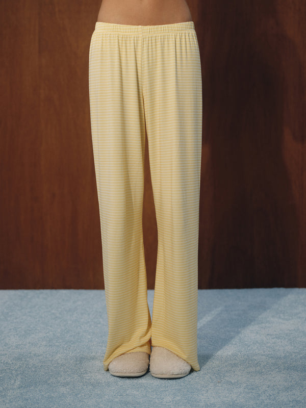 Yellow and white striped wide-leg lounge pants worn by a model, paired with beige slippers, standing on a blue carpet against a dark wood panel background.