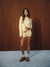 A woman stands against a dark wood-paneled wall wearing a matching cream two-piece lounge set with a cropped long-sleeve top and shorts, white socks, and brown slip-on shoes.