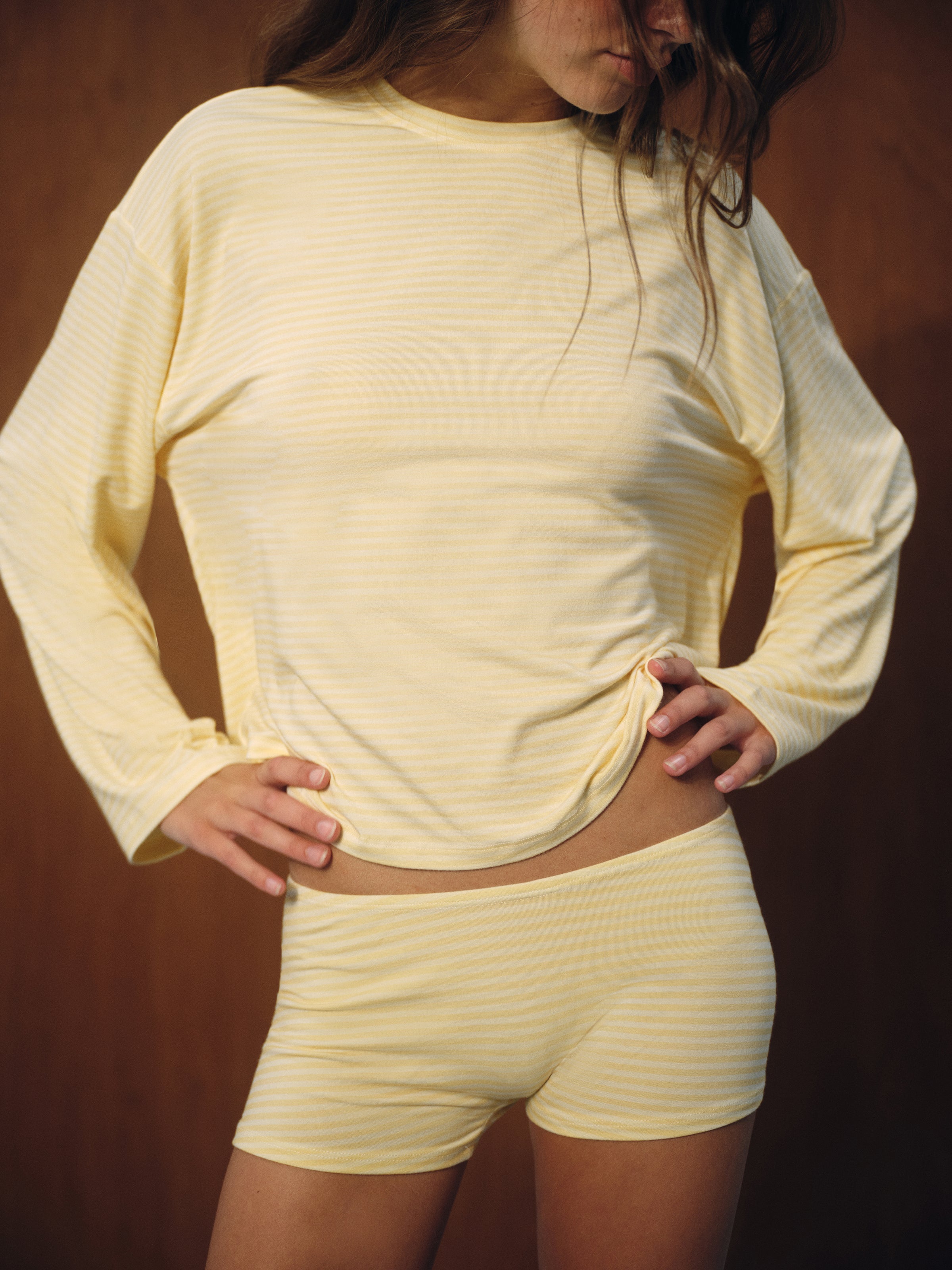 A woman wearing a pale yellow and white striped long-sleeve top and matching shorts, posing against a dark brown background. 
