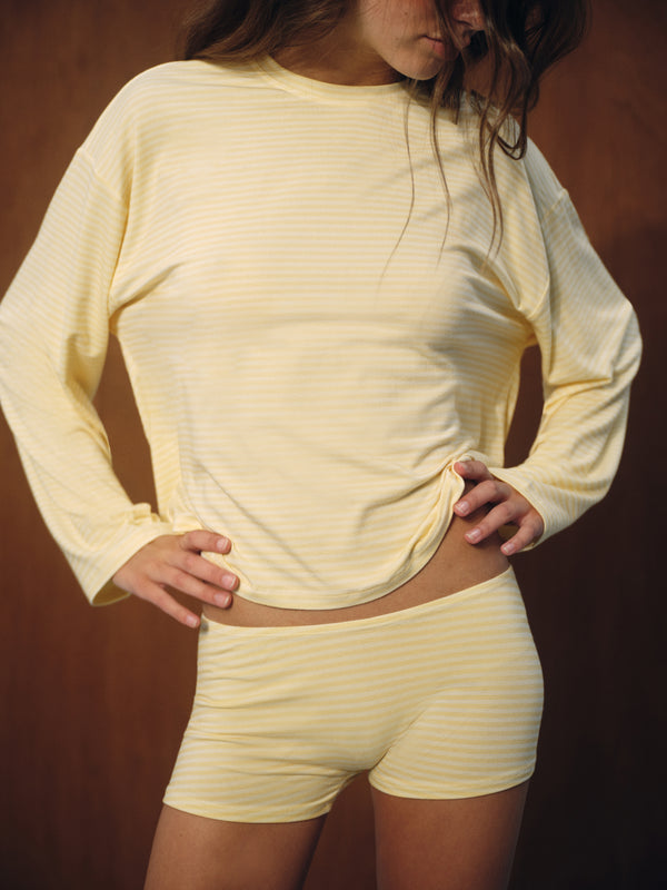 A woman wearing a pale yellow and white striped long-sleeve top and matching shorts, posing against a dark brown background. 