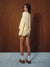 A woman stands in profile against a dark wood-paneled wall, wearing a pale yellow ribbed knit long-sleeve top and matching shorts, white socks, and brown slip-on shoes.