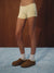 A person wearing pale yellow and white striped ribbed shorts, white socks, and brown slip-on slippers, standing on a blue carpet against a dark wood-paneled wall.