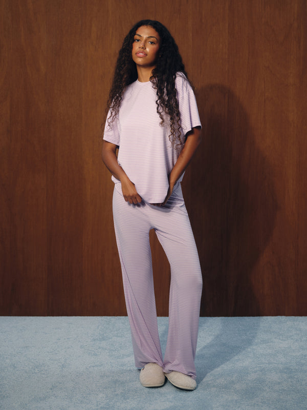 A woman modeling a light lavender ribbed lounge set consisting of a short-sleeve top and matching wide-leg pants, standing against a dark wood-paneled wall.