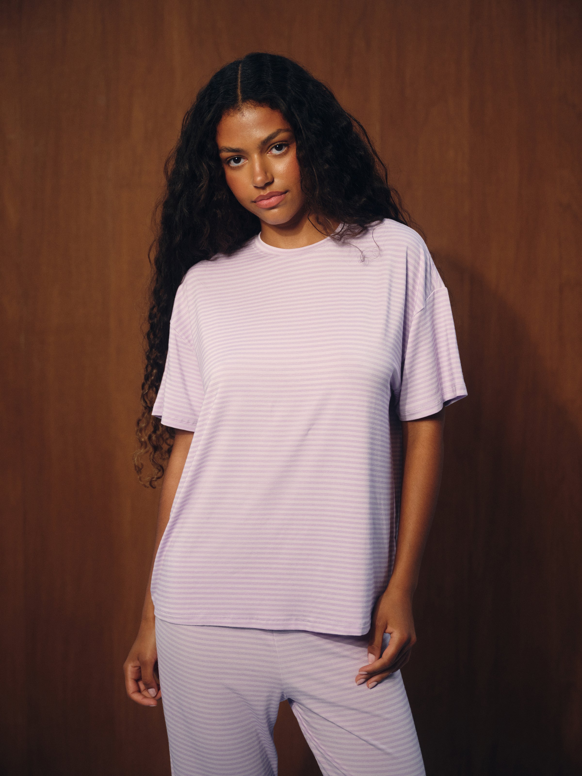 A woman with long, curly black hair wearing a light lavender short-sleeve t-shirt and matching striped lounge pants, standing against a dark brown wood-paneled background. 