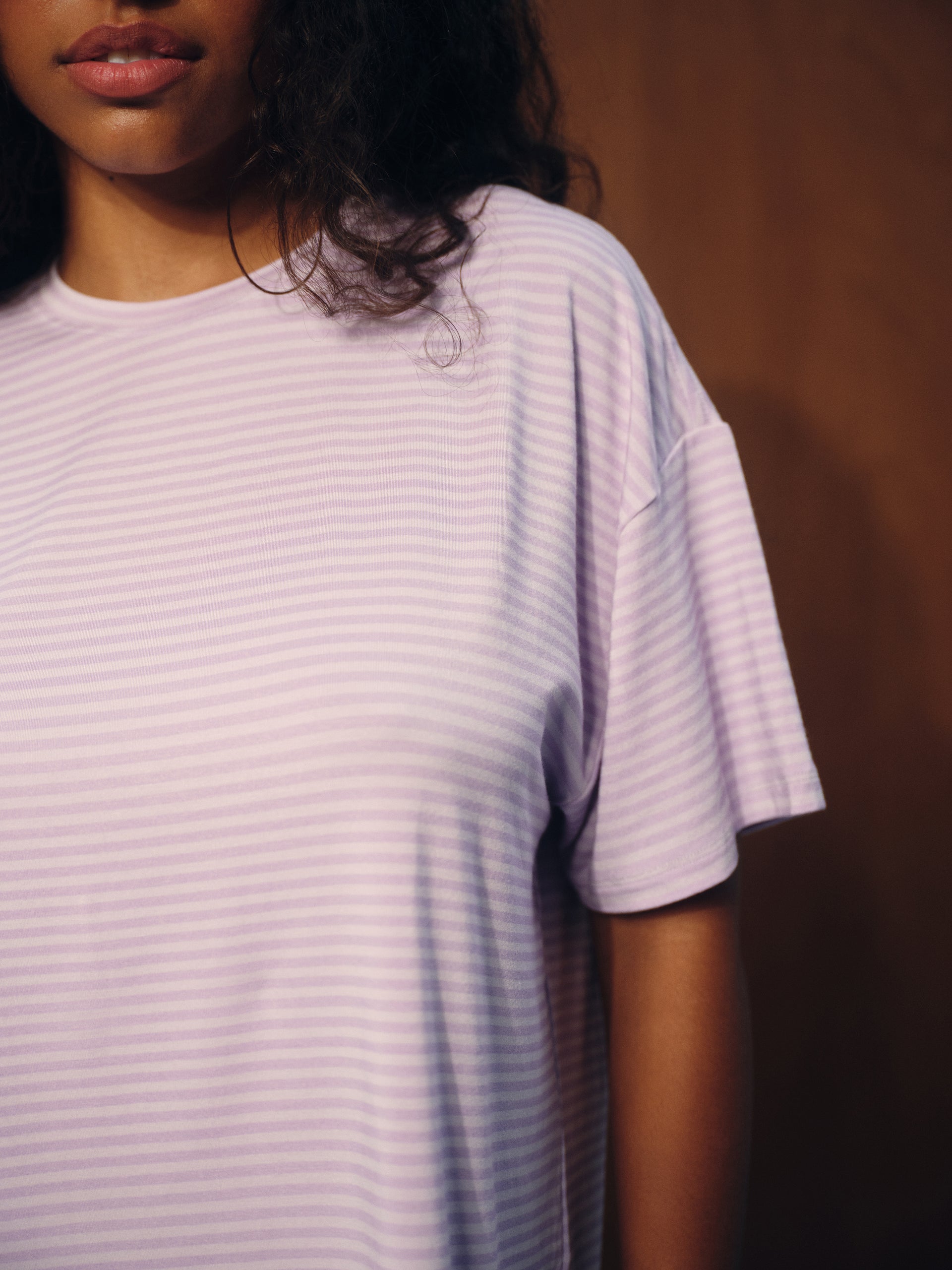 A close-up of a person wearing a short-sleeve crewneck t-shirt with fine horizontal lavender and white stripes, set against a dark brown background.