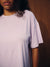 A close-up of a person wearing a short-sleeve crewneck t-shirt with fine horizontal lavender and white stripes, set against a dark brown background.