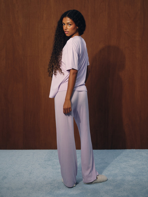 A woman stands in profile wearing a light lavender ribbed lounge set consisting of a short-sleeve top and wide-leg pants, set against a dark wood-paneled wall and a light blue-gray carpet.