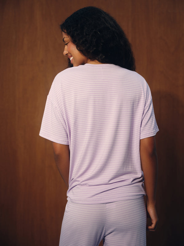 A woman shown from behind wearing a light lavender and white horizontally striped short-sleeve t-shirt and matching striped pants, standing against a dark brown wood-paneled background.