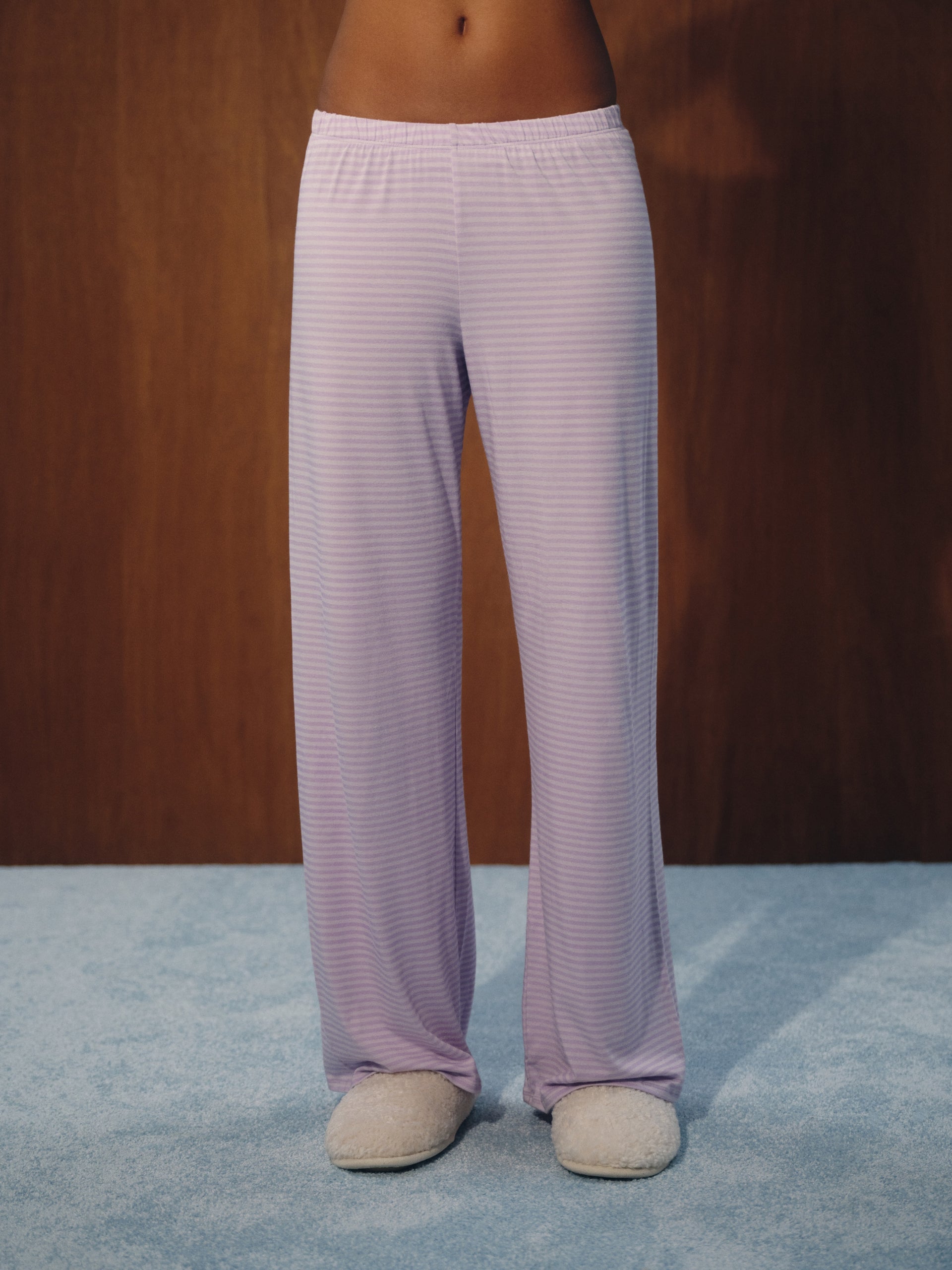 A person wearing light lavender striped lounge pants and fuzzy beige slippers, standing against a dark wood wall and blue carpet.