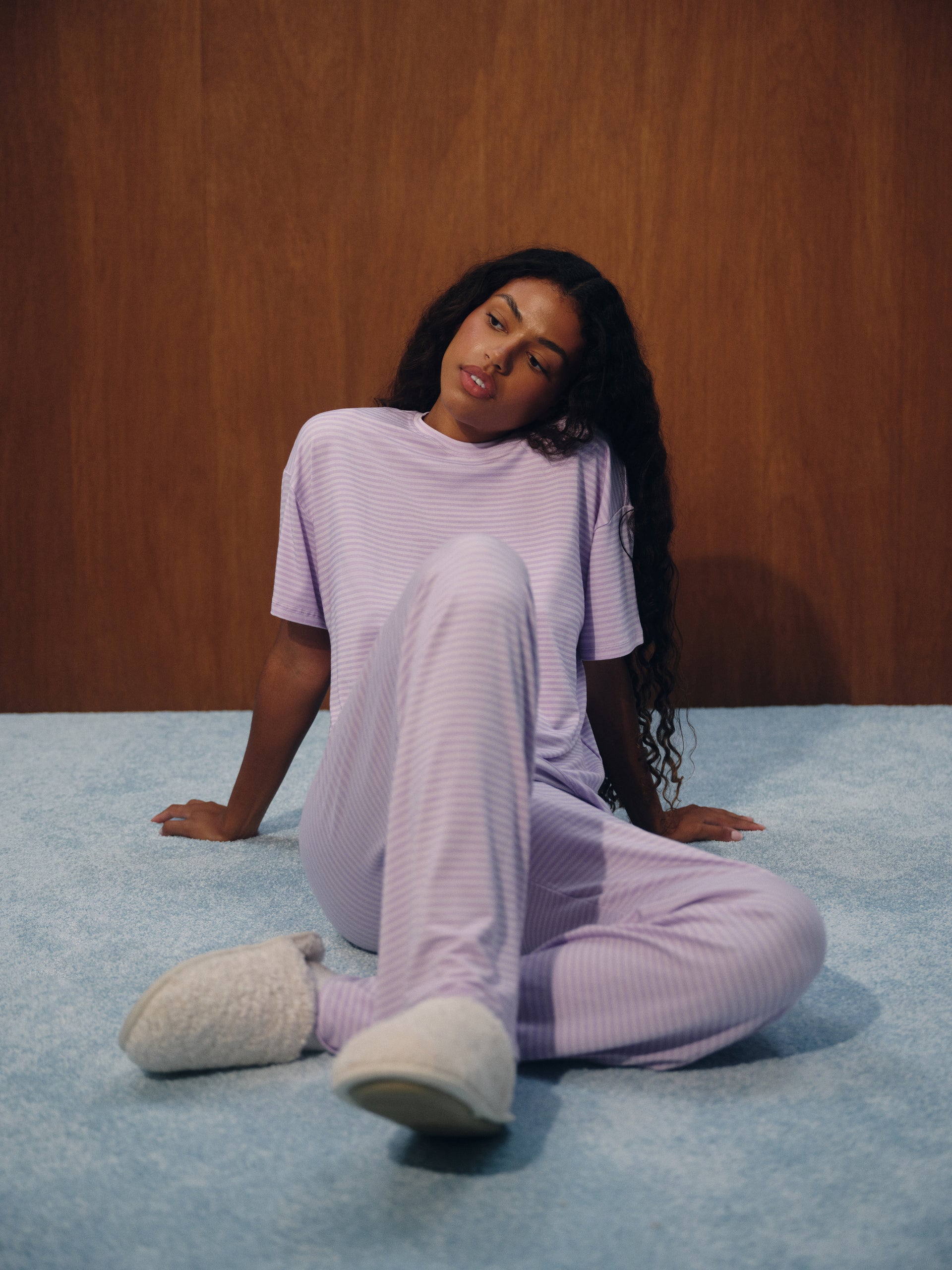 A woman sits on a light blue carpet in front of a wooden wall, wearing a light purple ribbed pajama set with matching fuzzy slippers.