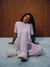 A woman sits on a light blue carpet in front of a wooden wall, wearing a light purple ribbed pajama set with matching fuzzy slippers.