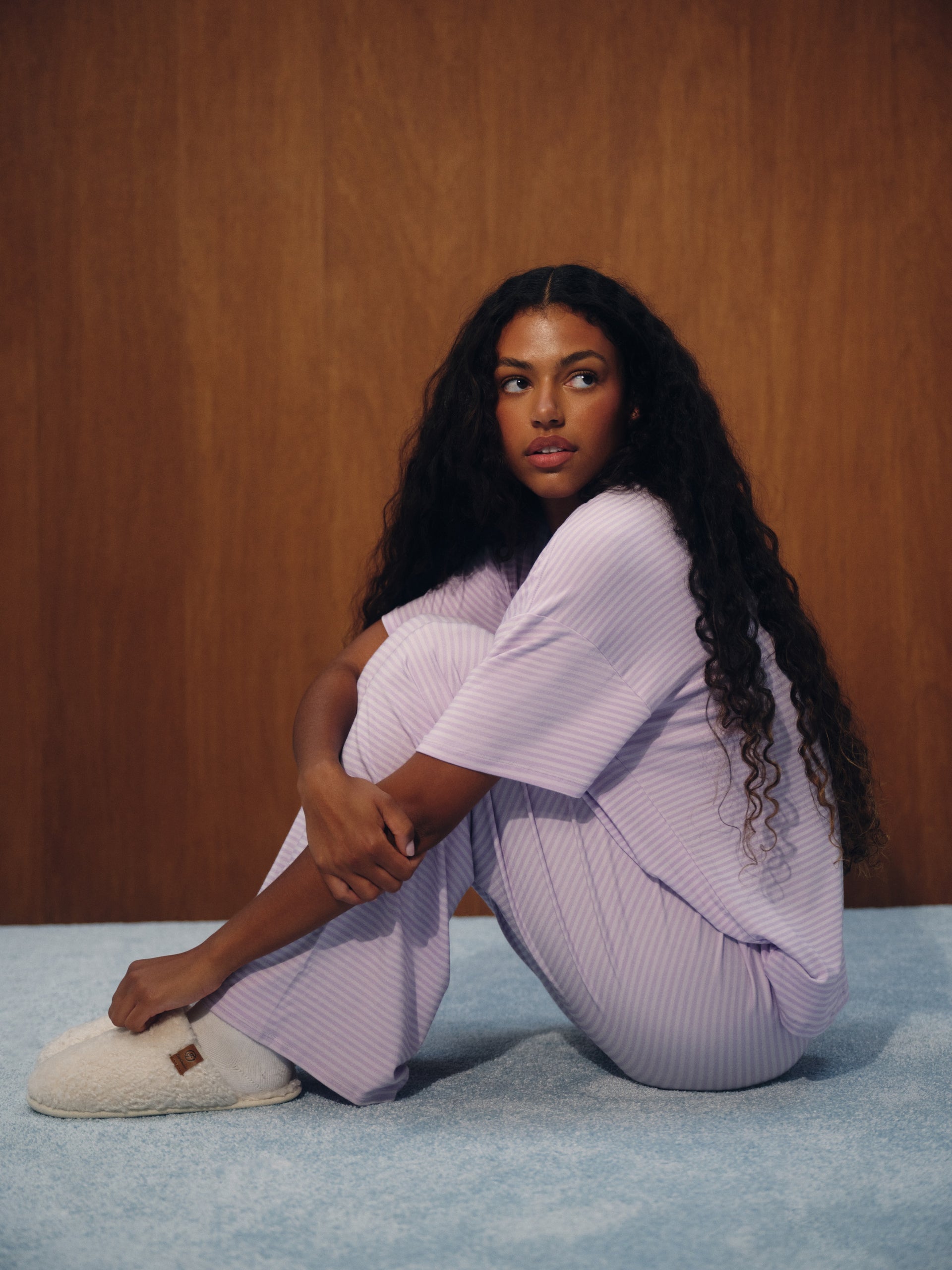 A woman with long, curly black hair sits on a light blue carpet in front of a brown wood-paneled wall, wearing a light purple/lavender ribbed pajama set and a white slipper.