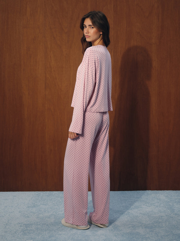 A woman wearing a light pink two-piece pajama set with a small white polka dot pattern, standing in profile against a dark wood-paneled wall.