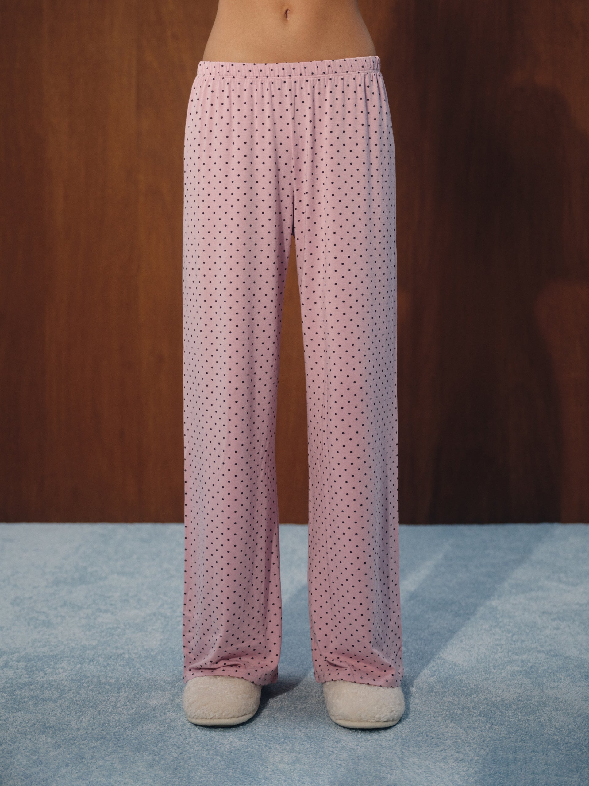 Pink wide-leg pajama pants with a small dark polka dot pattern, shown on a model from the waist down against a dark wood background and blue carpet.