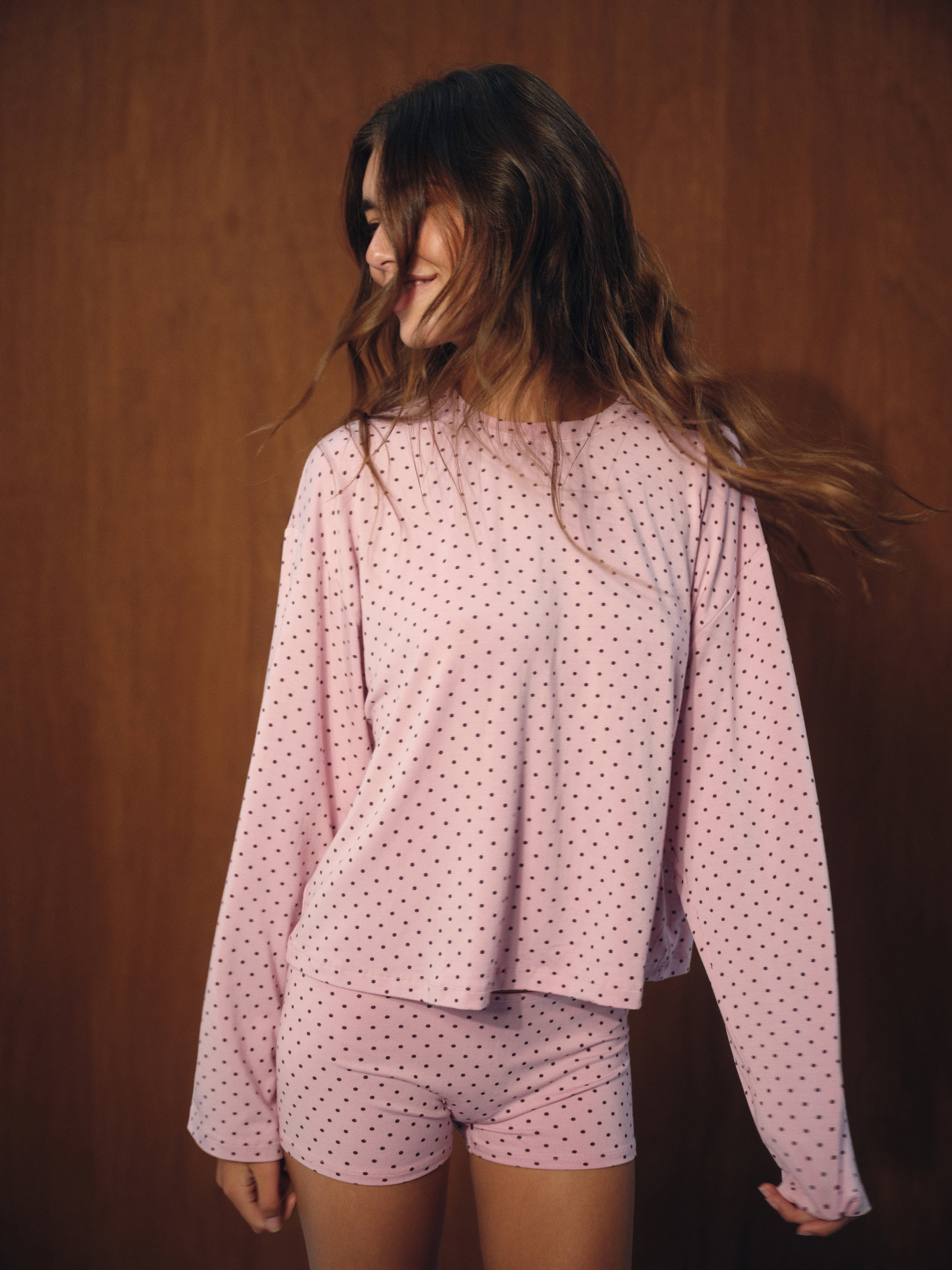 A woman wearing a light pink long-sleeve pajama top and matching shorts with a small dark polka dot pattern, posed against a dark brown wooden background.  