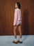 A woman stands in profile wearing a light pink long-sleeve top and matching shorts with a small white polka dot pattern, paired with white socks and brown slip-on shoes, set against a dark wood-paneled wall and a light blue floor.