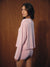 A woman wearing a light pink two-piece pajama set with small dark polka dots, standing against a dark wood-paneled background.