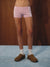 A person from the waist down wearing light pink boxer briefs with a small dark polka dot pattern, white socks, and brown slip-on shoes, standing against a dark wood panel background.