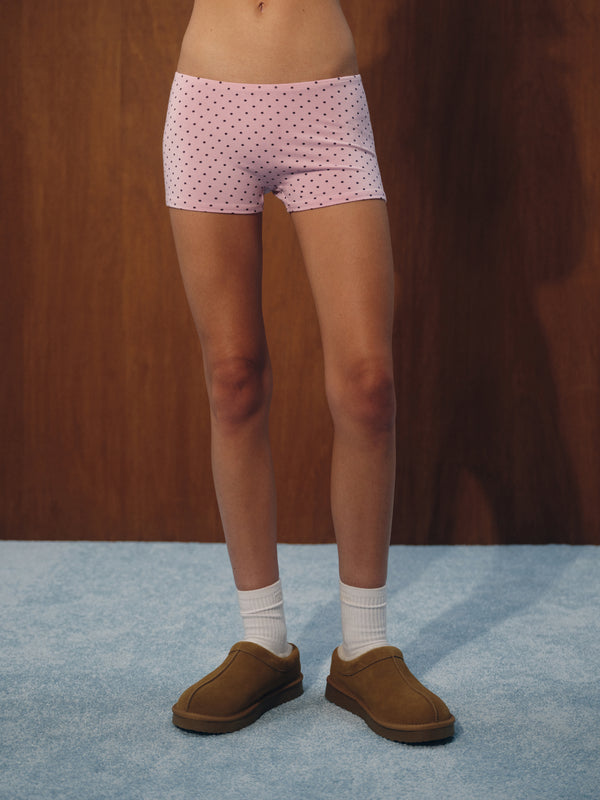 A person from the waist down wearing light pink boxer briefs with a small dark polka dot pattern, white socks, and brown slip-on shoes, standing against a dark wood panel background.