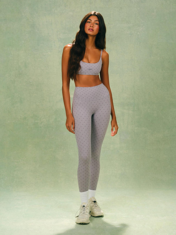 A woman modeling a matching light gray two-piece activewear set with a subtle polka dot pattern, consisting of a sports bra and high-waisted leggings, paired with white socks and white sneakers, set against a textured sage green studio backdrop. 