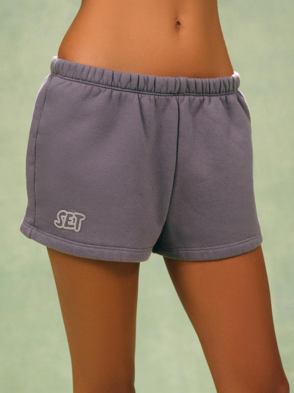 A person wearing gray athletic shorts with an elastic waistband and a white embroidered 'SET' logo on the lower left leg, shown against a soft green background. 