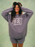 A woman smiling and laughing while wearing an oversized lavender-gray sweatshirt with a large white 'SET' graphic on the front, paired with matching shorts, against a textured sage green background. <model-info>Meeya wears a size XS/S and is 5’7, 23” waist, 32B bust.</model-info> 