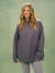 A woman modeling an oversized dark gray sweatshirt with a small embroidered logo on the left chest, paired with gray polka-dot leggings, against a textured sage green studio backdrop. <model-info>Raine wears a size L and is 5’8”, 29½” waist, 34” bust.</model-info>
