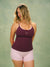 A woman modeling a matching sleepwear set consisting of a dark plum purple tank top with a small white 'SET' logo and light pink polka-dot shorts, against a textured sage green  <model-info>Raine wears a size L and is 5’8”, 29½” waist, 34” bust.</model-info>background.
