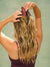 A woman with long, wavy blonde hair seen from behind, wearing a burgundy hair claw clip with the brand name 'SET' visible on the strap, against a muted sage green background.