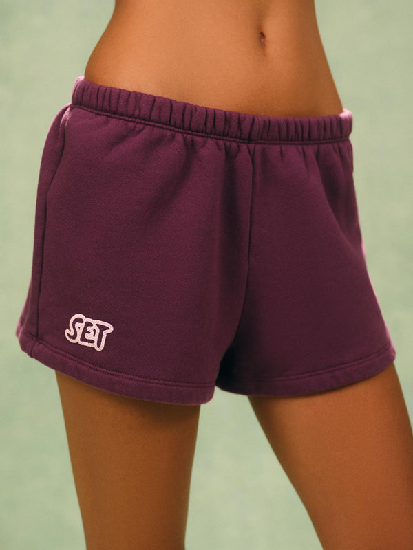 A close-up of a person wearing dark purple athletic shorts with an elastic waistband and a white embroidered 'SET' logo on the left leg, set against a soft green background. 