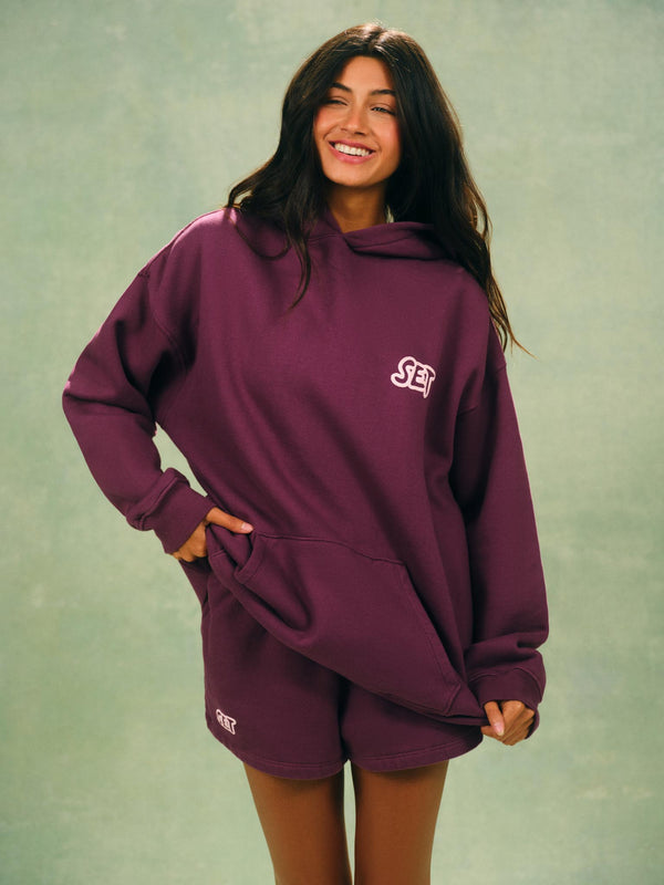 A woman modeling an oversized maroon hoodie and matching shorts set with a small white embroidered logo on the chest and leg, set against a textured sage green background. 