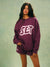 A woman modeling an oversized maroon sweatshirt and matching shorts set with a white 'SET' logo on the front, posed against a textured sage green background.