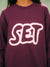 A close-up of a person wearing a crewneck sweatshirt in deep plum purple with a large white graphic spelling 'SET' across the chest, decorated with tiny pink dots.