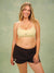 A woman modeling a light yellow and black polka dot sports bra and dark brown shorts against a textured green studio backdrop. <model-info>Raine wears a size L and is 5’8”, 29½” waist, 34” bust.</model-info>