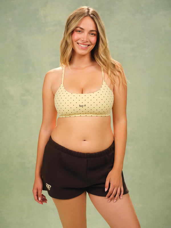 A woman modeling a light yellow and black polka dot sports bra and dark brown shorts against a textured green studio backdrop. 