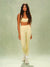 A woman modeling a matching pale yellow two-piece activewear set with small black polka dots, consisting of a cropped tank top and high-waisted leggings, paired with white sneakers against a textured sage green studio backdrop.