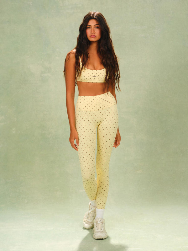A woman modeling a matching pale yellow two-piece activewear set with small black polka dots, consisting of a cropped tank top and high-waisted leggings, paired with white sneakers against a textured sage green studio backdrop.