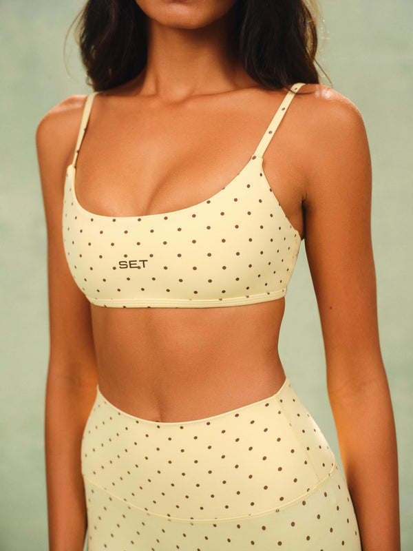 A woman wearing a matching cream-colored activewear set with brown polka dots, featuring a sports bra and high-waisted leggings, against a soft sage green background. 