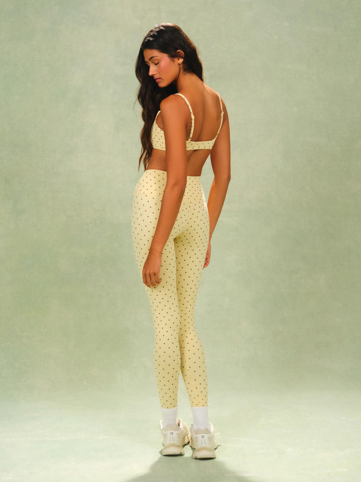 A woman stands in a studio wearing a matching pale yellow activewear set with small dark polka dots, consisting of a sports bra and high-waisted leggings, paired with white socks and white sneakers against a soft green backdrop.