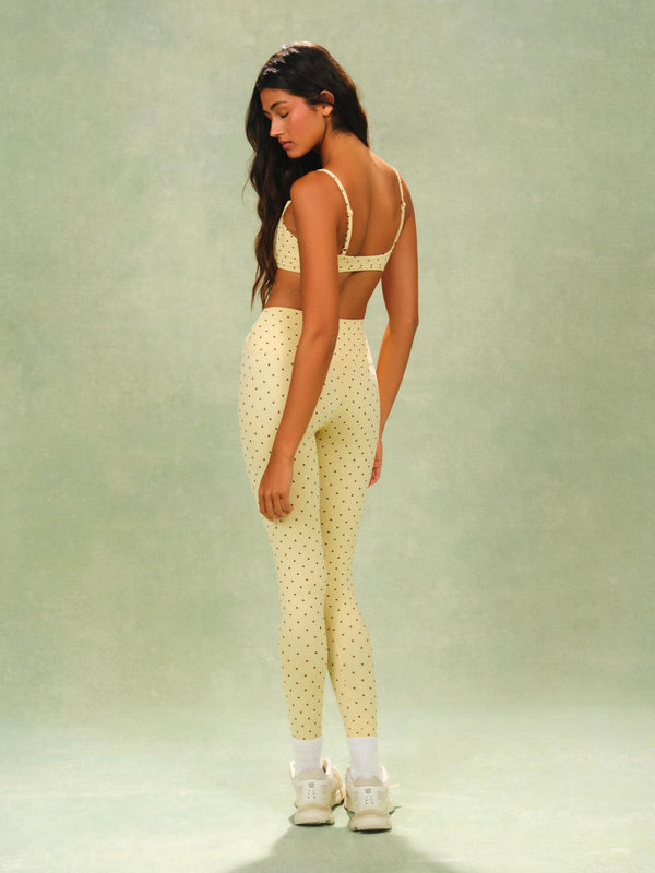 A woman stands in a studio wearing a matching pale yellow activewear set with small dark polka dots, consisting of a sports bra and high-waisted leggings, paired with white socks and white sneakers against a soft green backdrop.
