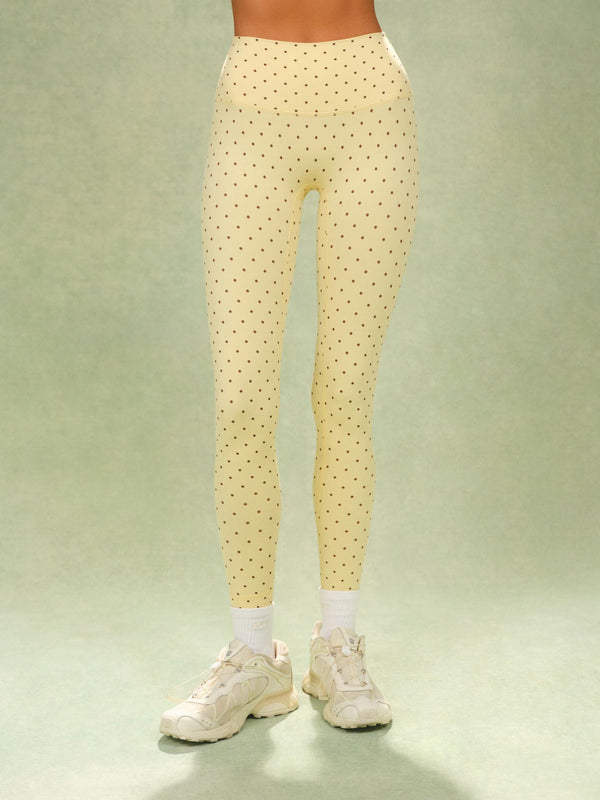 A person from the waist down wearing high-waisted, light yellow leggings with a small black polka dot pattern, styled with white socks and cream-colored athletic sneakers against a light green studio backdrop.