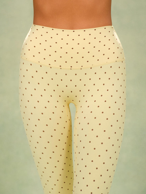 Close-up of a model wearing high-waisted leggings in a pale cream color with a small brown polka dot pattern, set against a solid sage green background.
