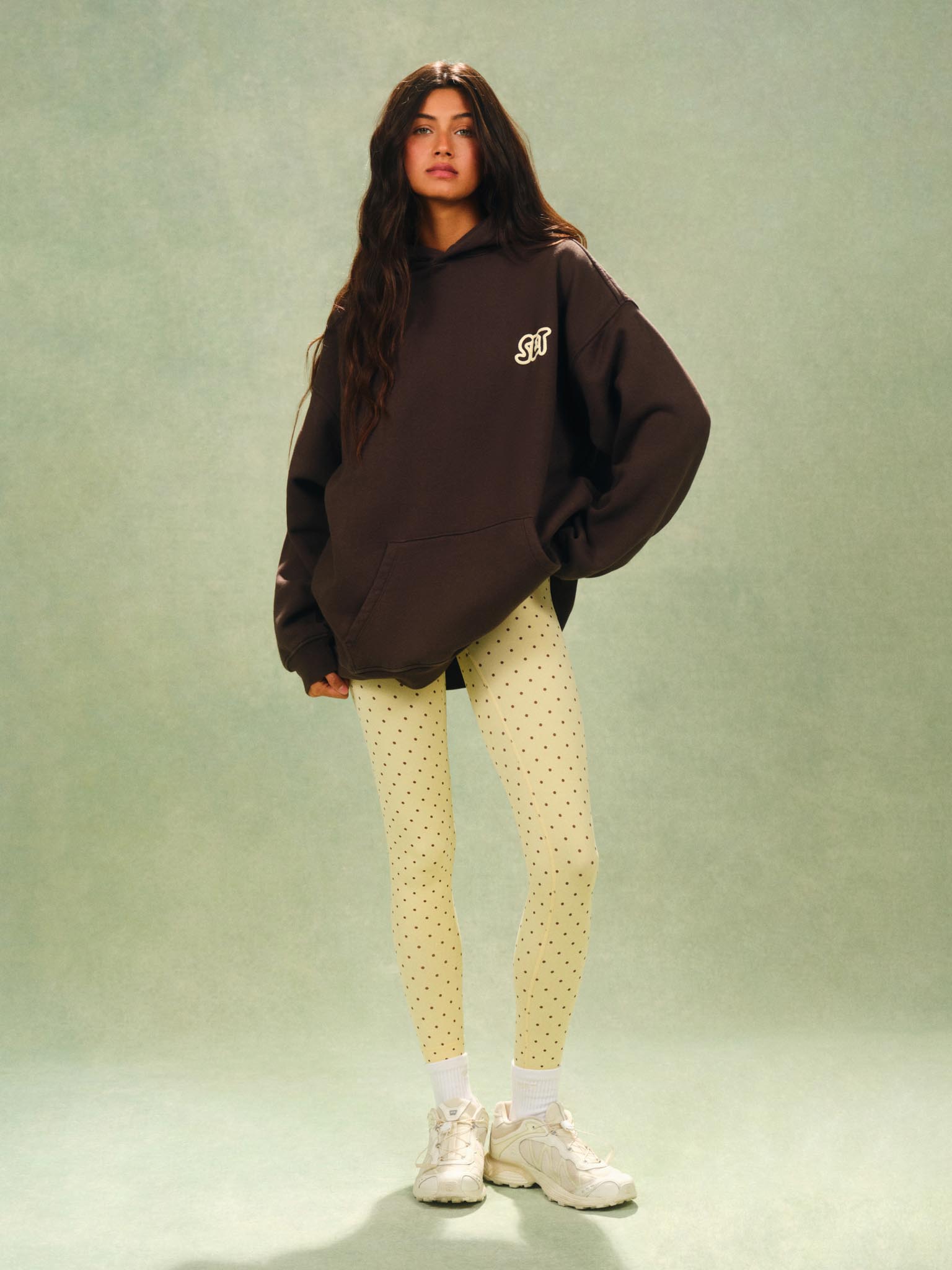 A woman stands against a muted sage green studio backdrop wearing an oversized dark brown hoodie with a small white chest graphic, pale yellow dotted leggings, white socks, and chunky white sneakers.