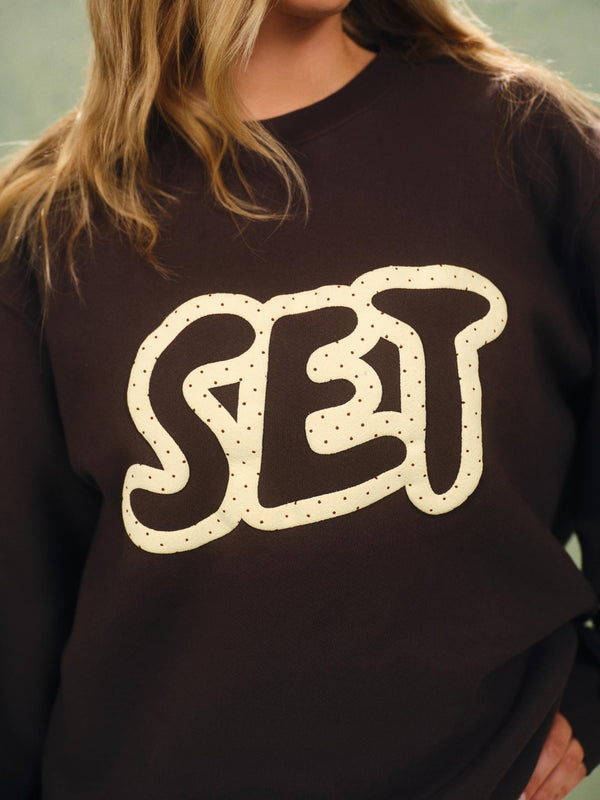 A close-up of a person wearing a dark brown crewneck sweatshirt with a large cream-colored graphic spelling 'SET' outlined with small dots.